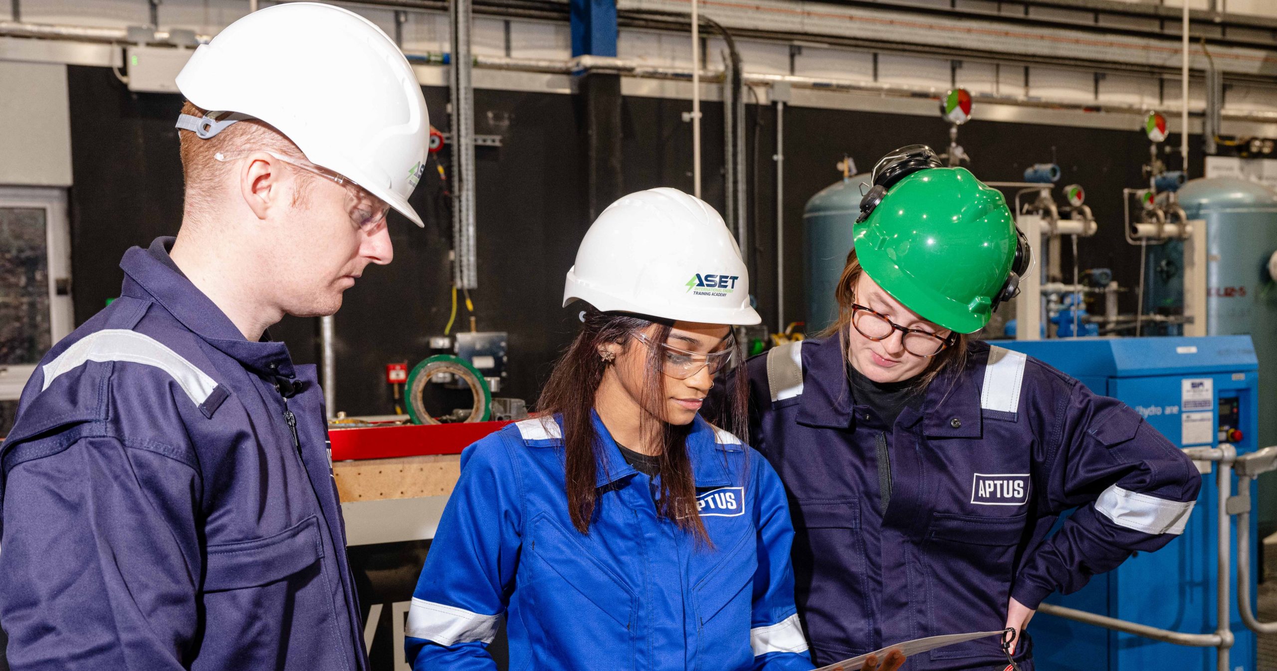 Three apprentices in an industrial training facility examine a document together.