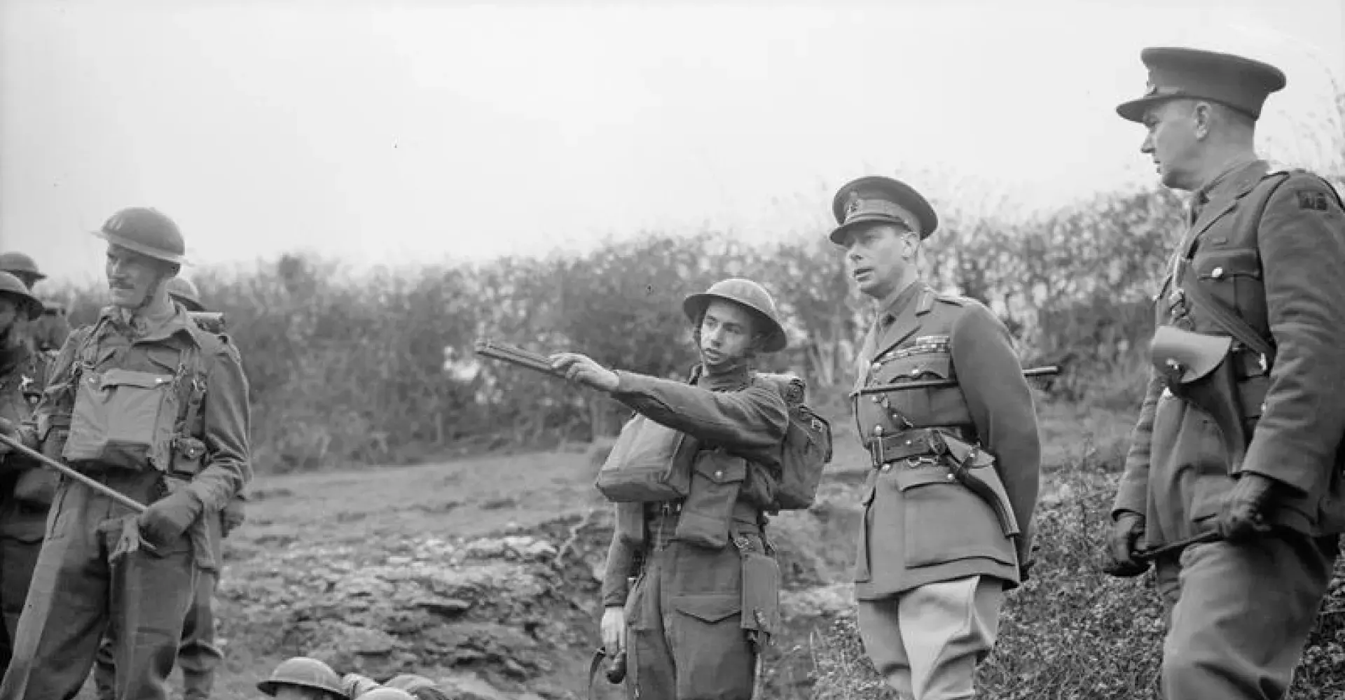 King George VI stands with troops watching a manoeuvre, which is not pictured. The soldier to his right is pointing to the manoeuvre in the distance. 