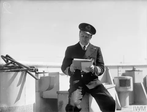 Lieut Cdr F C V Brightman sits on a box on the deck of a ship reading a manuscript