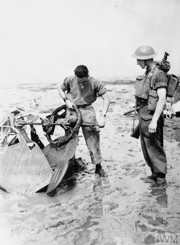 Historic B&W photograph of two soldiers standing in mud on the foreshore examine aircraft wreckage.