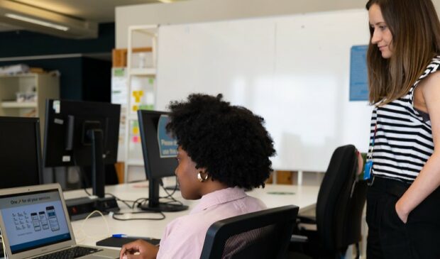Two people looking at designs on a screen in the GDS London office. One is sitting with a laptop and the other is standing behind.