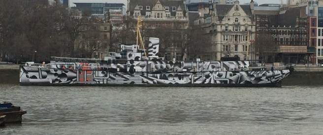 Photograph of HMS President moored on river in predominantly black and white dazzle camouflage scheme, with some red, against a backdrop of buildings on the river bank.