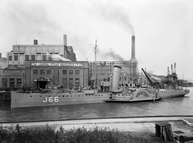 Black and white photograph of paddle steamer marked with pennant number J66 to the left, with its funnel echoed in the chimneys of the industrial buildings beyond.