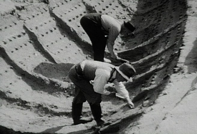 Historic 1939 B&W film still showing archaeologists at work within the 'ship-shape' with the ship structure visible in the sand, and all the rivets still in situ.