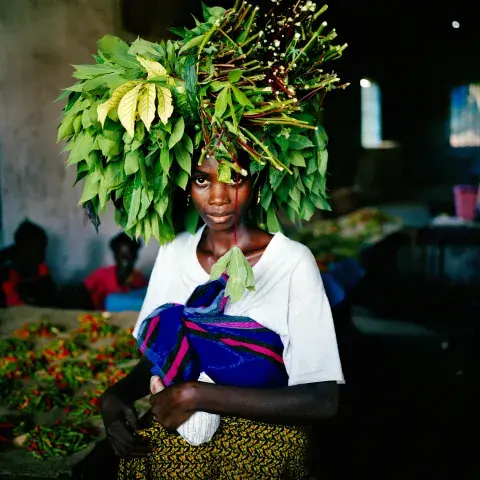 Liberian woman looking at the camera, carrying cassava leaves on her head to central market in Tubmanburg during Second Liberian Civil War, May 2003.