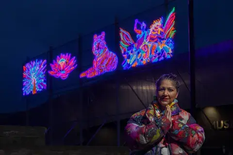 Chila Burman in front of IWM North at night. Neon artworks created by Chila are on display behind her.
