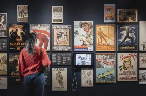 Back of female visitor looking on at exhibition wall of objects containing selection of propaganda posters. 