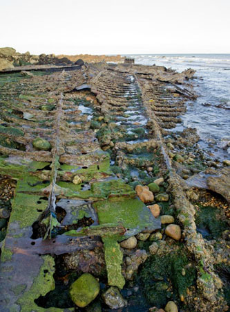 Portrait format photograph showing all that remains of the burnt-out Falcon, with the bottom ribs infilled with stones and green seaweed, sea lapping at the upper right-hand edge of the photograph.