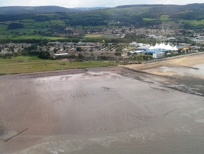 Aerial view of beach with small wreck visible at centre right, with green fields and hills beyond.