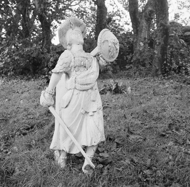 B&W photo of white-painted figurehead in Scottish dress with tam o'shanter, kilt, sword and shield, in churchyard.