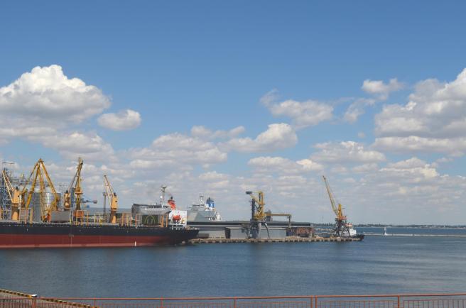 Dockyard with cranes to left, sea to right, against a blue sky with fluffy clouds.