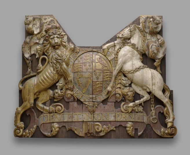 Carved wooden relief panel with some faded colour, showing 17th century royal shield of arms in centre, with lion supporter on left and unicorn supporter on right.