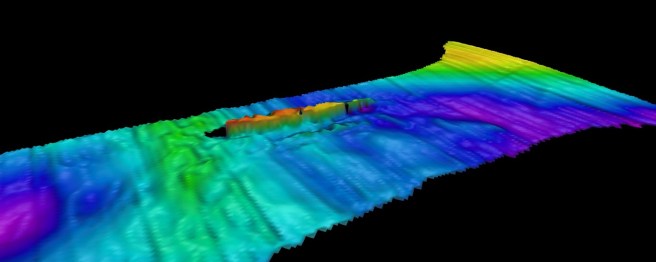 Multi-coloured textured image of seabed set against a plain black background. The seabed is primarily greens and blues, with a raised section in reddish hues representing the wreck above the seabed.
