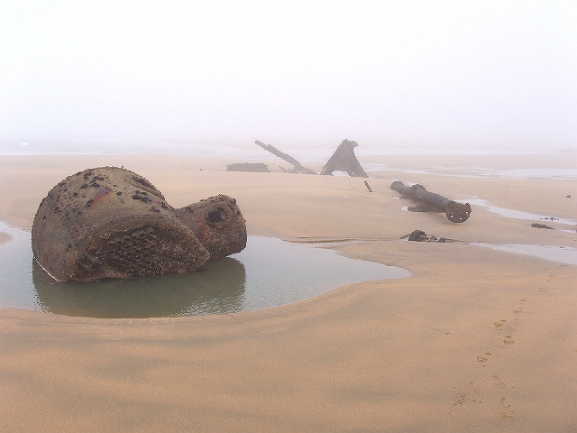 Three groups of dark metal elements of a wreck sitting in pools of water on a sandy beach, against a backdrop of grey mist.