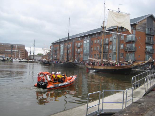 View of Gloucester Docks with a series of tall ships, and an orange search and rescue vessel in foreground.