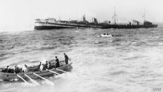 Black and white photograph of rowing boats in foreground and middle ground, steering away from sinking hospital ship in the background. To the right background a black vessel stands by to assist.