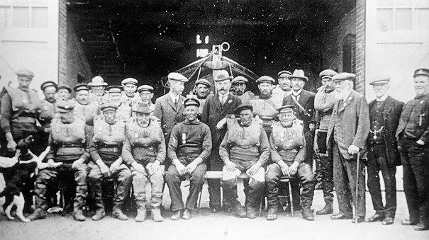 Black and white photograph of two rows of en, seated in front, standing at the back, in front of the open doorway through which the bows of a lifeboat can be seen.