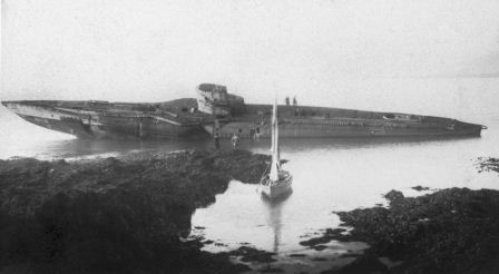 Black and white photo of a wrecked submarine, which bisects the photograph longitudinally. In the foreground dark rocks and an inlet, in which the white sail of a dinghy can be seen. The background grey, featureless sea and sky.