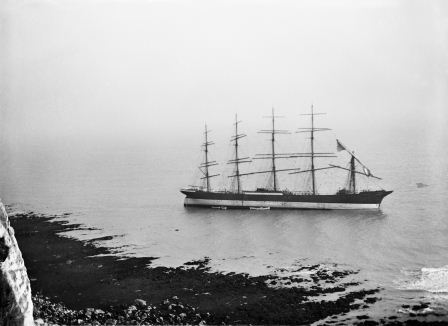 Black and white photograph of five-masted sailing ship aground in shallow water off a rocky coastline in the foreground. 