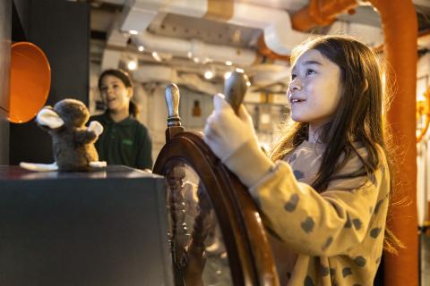 A young girl "steers" HMS Belfast, moving the ship's wheel.
