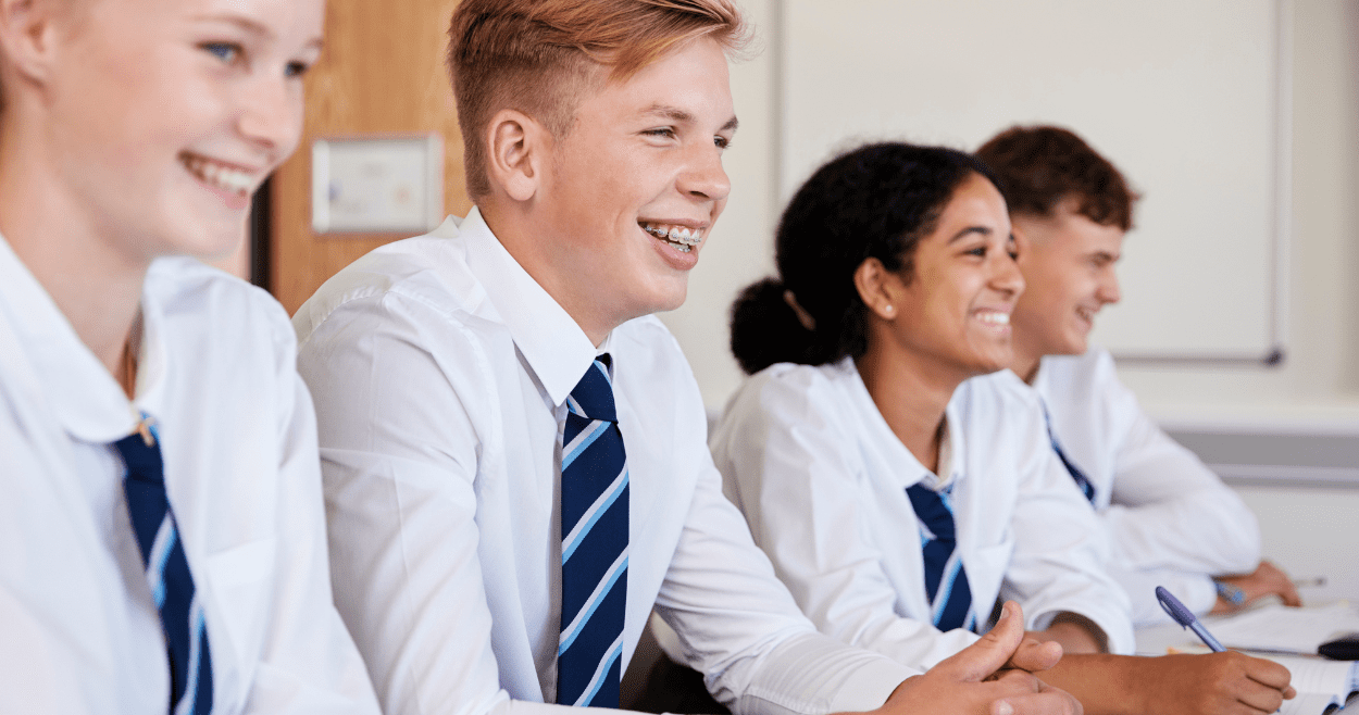 Four secondary school students smiling at a desk wearing school uniform