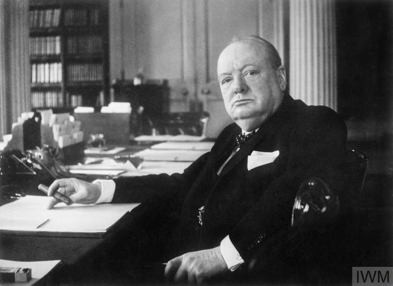 Winston Churchill sits at a desk, his body slightly angled towards the camera.
