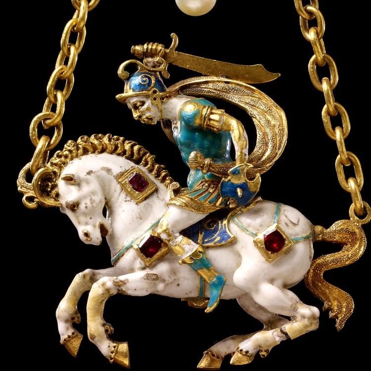 Close-up view of a bejewelled pendant in the form of a warrior holding up a sword while riding a white horse, suspended from the head and tail by a gold chain.
