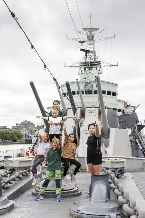 A group of children stand in front of HMS Belfast's turret, pointing at the camera.