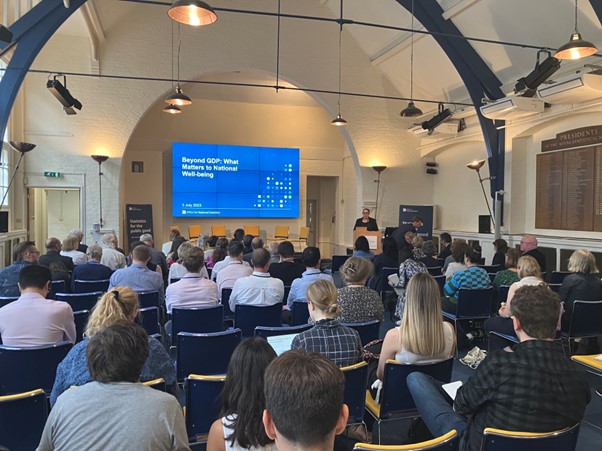 A presentation being given to a crowded room, titled "Beyond GDP: What Matters to National Well-being"