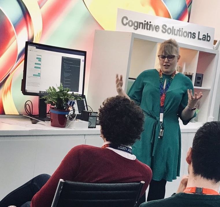 A female presenter is standing talking to her audience in a brightly coloured high tech office