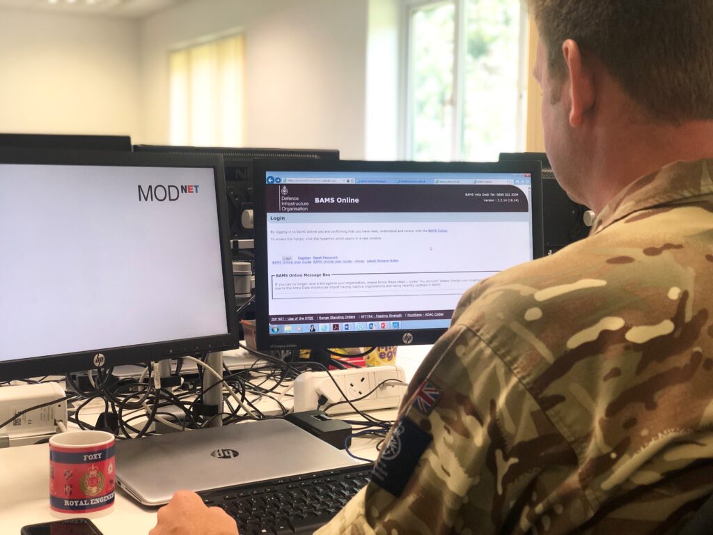Image of service personnel wearing his army uniform facing two computer screens. One computer screen on the right-hand side shows the BAMS online website and the left-hand side screen shows a blank white screen with MOD net on the top right-hand corner. 
