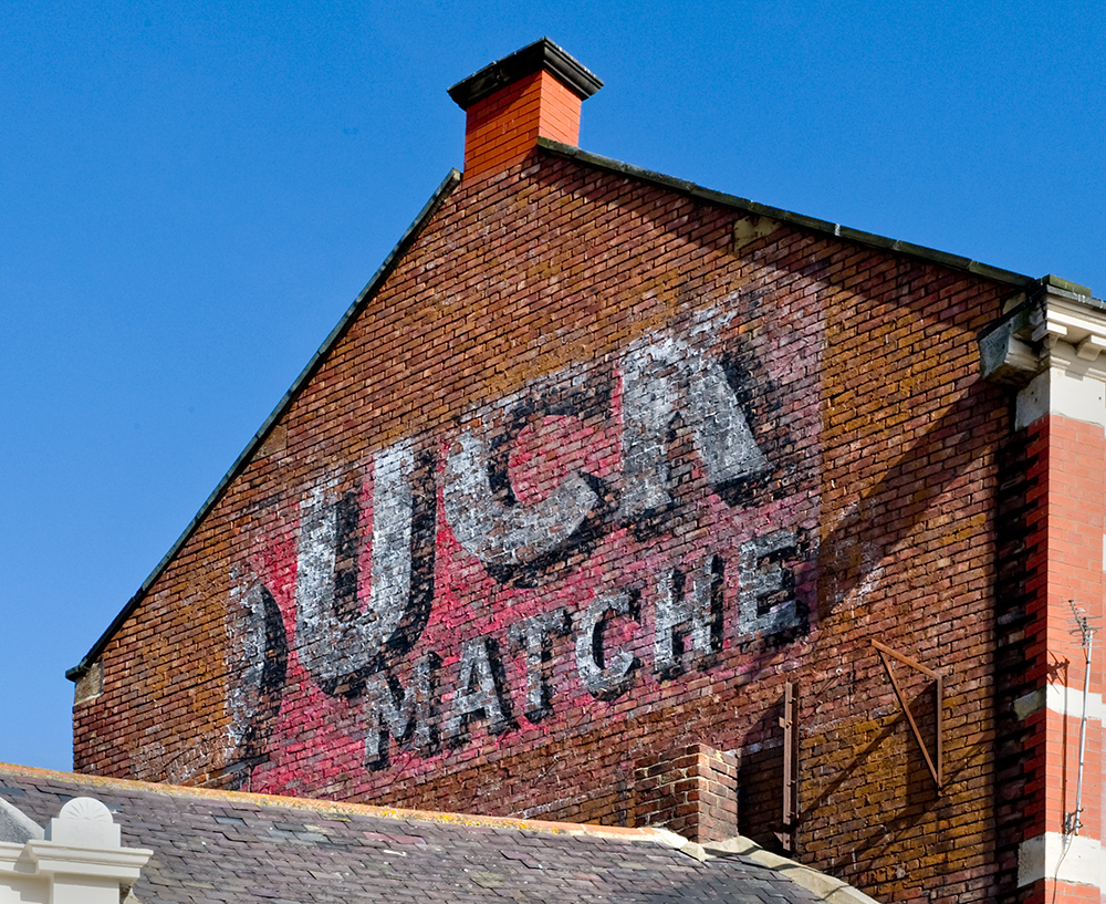 Image of the side of a brick building with a red faded advertising sign for matches.