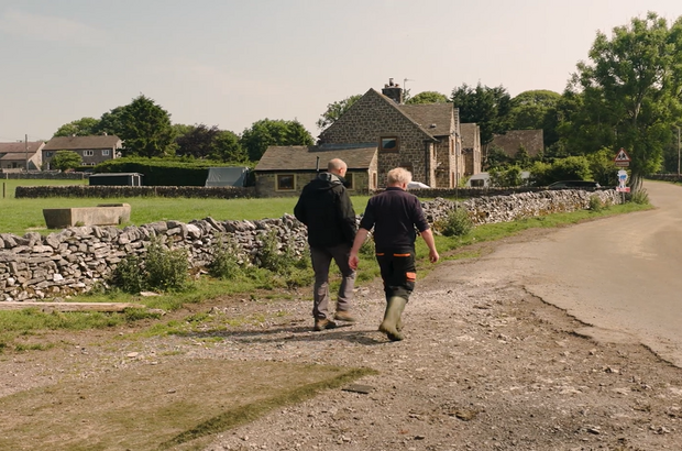 Two people walk away from the camera along a gravel road beside a dry-stone wall in a rural village. Stone houses and green fields sit in the background, with trees lining the road under a clear, sunny sky.