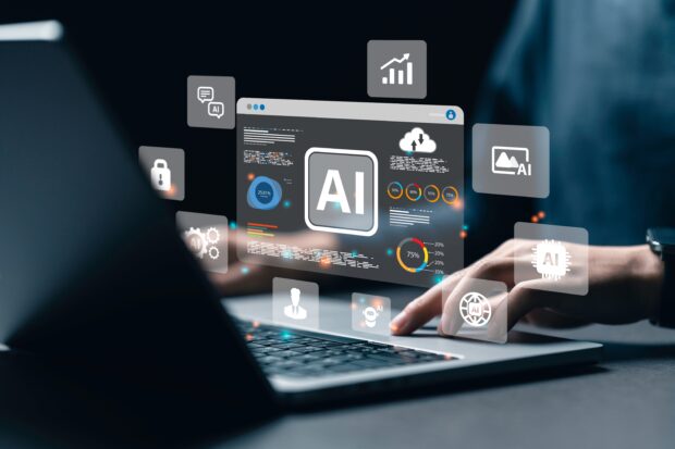 A person typing on a laptop with holographic icons related to artificial intelligence (AI) appearing above the keyboard. The central icon displays 'AI' prominently, surrounded by other icons including a bar chart, a lock, a speech bubble, a cloud with connected dots, and a human profile. The background is dark, highlighting the bright holographic elements.
