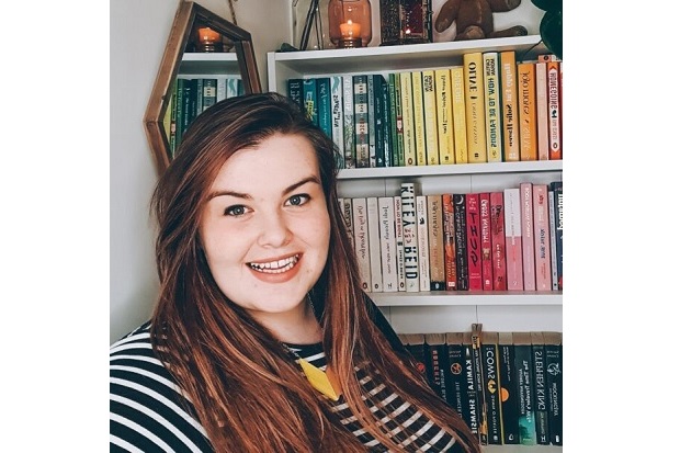 Alice sits smiling in front of a bookcase full of colourful books. She is wearing a top with black and white horizontal stripes and her long brown hair is worn down and parted to the side.