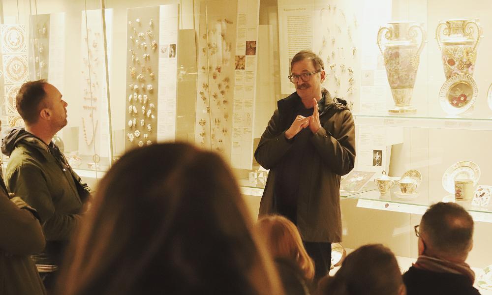 A guide standing in front of a glass Museum case filled with porcelain objects signs in BSL to a tour group.   