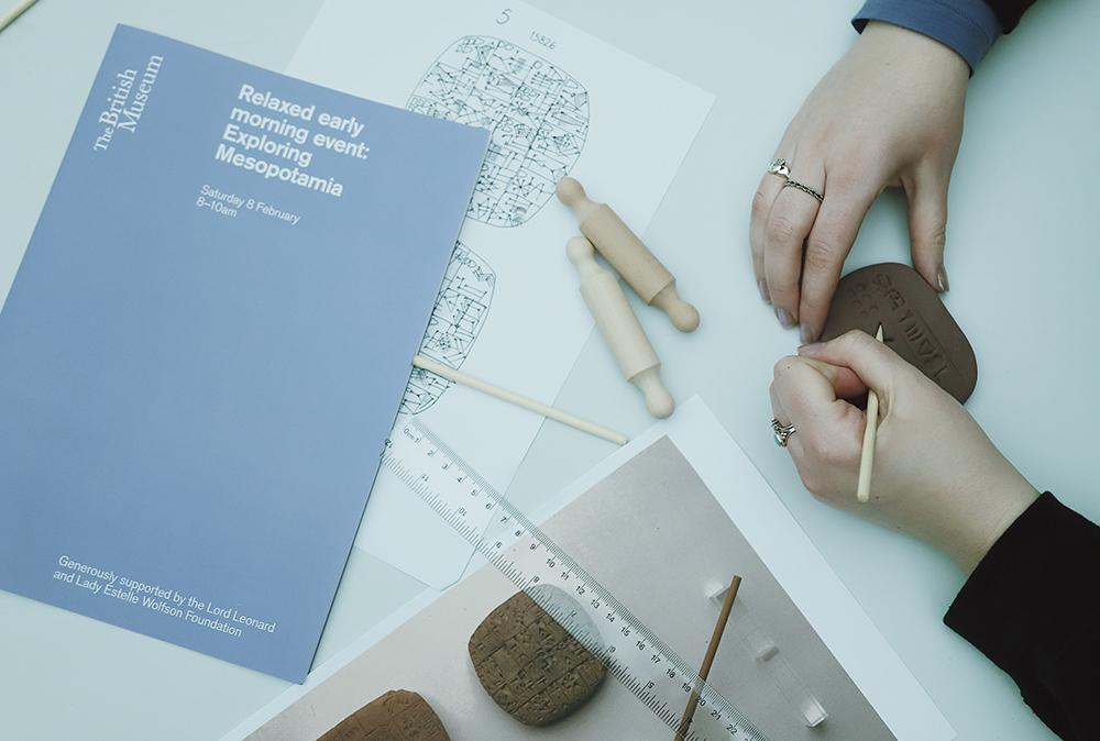 View of a table from above, with a paper event programme, two miniature rolling pins, a ruler and the hands of a person writing on a clay tablet with a wooden stick.  