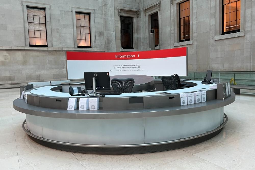 The round information desk in an open space near a corner of the Great Court, with five windows in the background. 