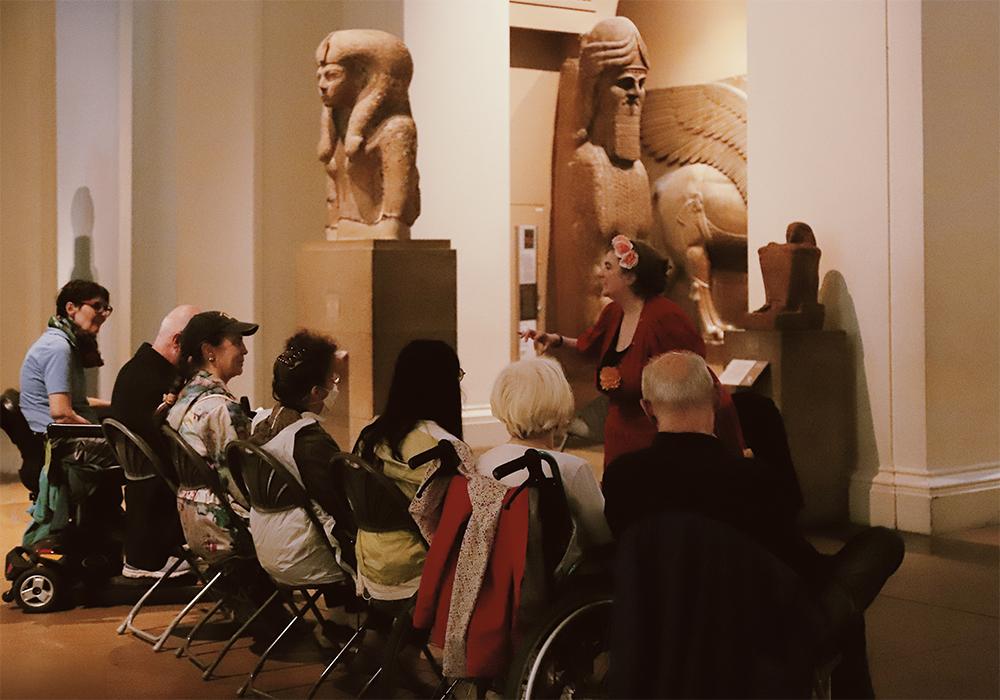 A tour guide talks to a group of seven seated people – one is in a wheelchair, and another is on a mobility scooter.  