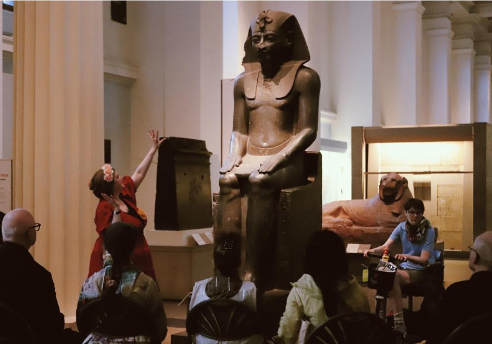 A tour guide talks to a group of six seated people – one is on a mobility scooter. 