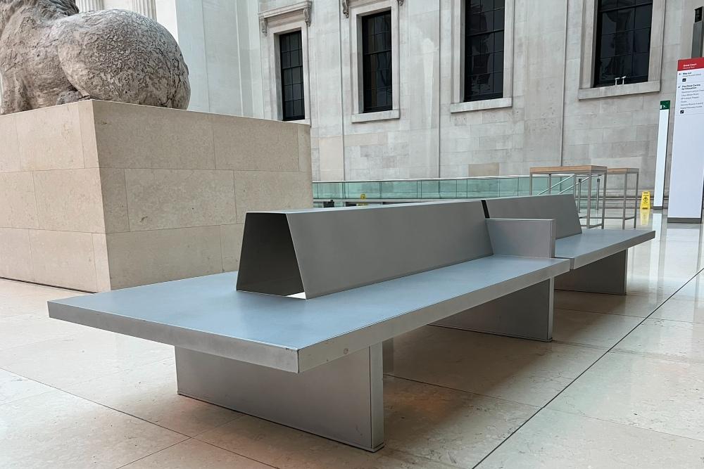 A long metal bench with a backrest and seating on either side. The back of a lion statue is visible in the top left and there are four windows on the right.