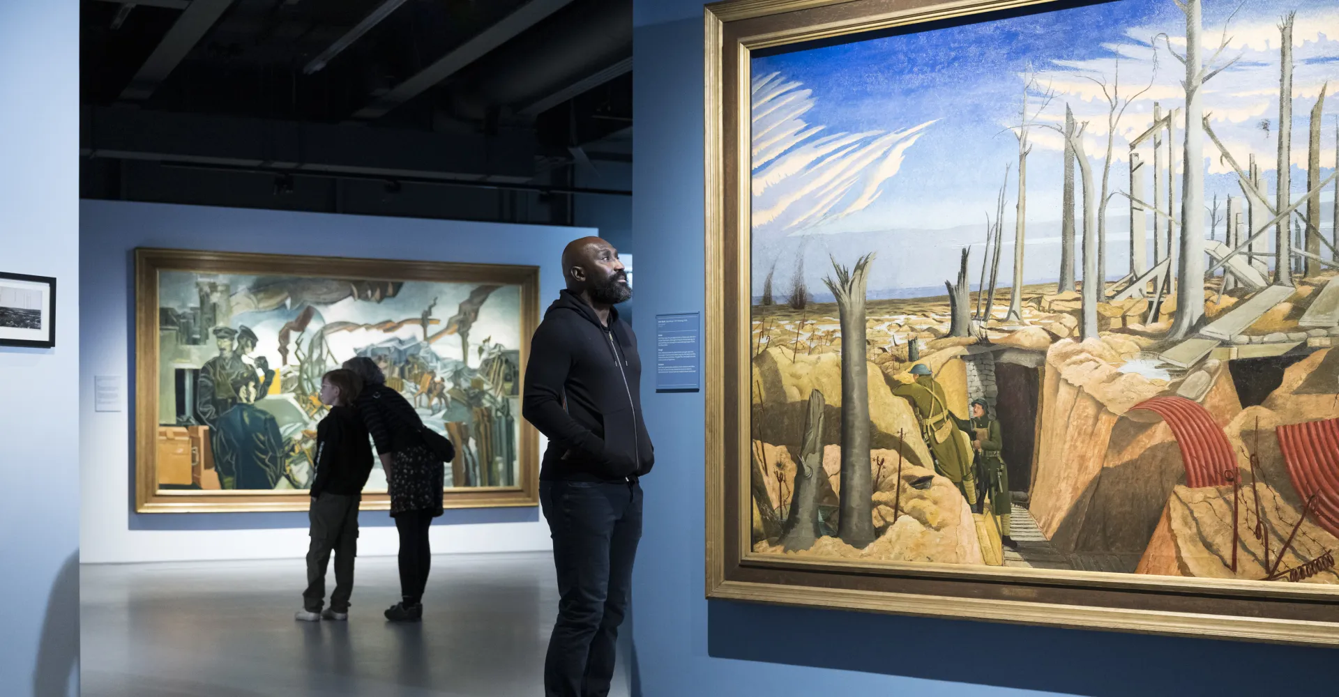 Visitors stood in IWM London's Blavatnik Art, Film and Photography galleries, admiring artworks, including 'Oppy Wood, 1917, Evening' by John Nash.