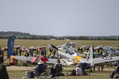 Spectators, staff and aircraft at Duxford airfield. Some visitors are up close looking at aircraft talking with staff.