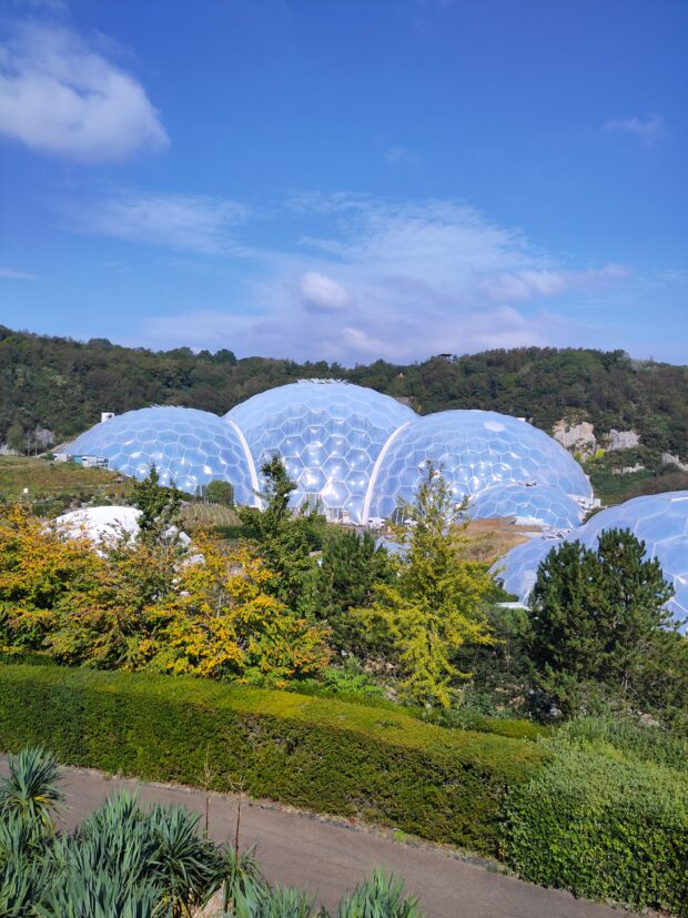 Image shows a picture of The Eden Centre, a blue sky hangs overhead with fluffy clouds. Lots of green trees are in the foreground, their leaves are just starting to change colour, indicating that it's likely later summer/early autumn time