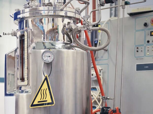 Stainless steel fermentation tank with pressure gauge, pipes and heat warning sign in a lab setting.