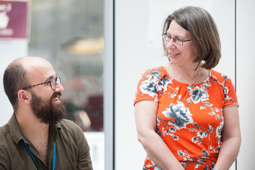 A seated male with a beard is smiling at a smiling female standing next to him