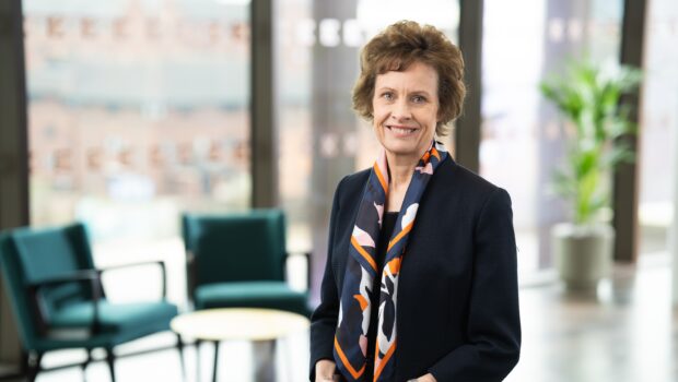 Professional portrait of a person standing indoors in a modern office setting. The person is wearing a dark blazer and a patterned scarf, facing forward. In the background, there are blurred chairs, a round table, large windows, and a potted plant, with soft natural light illuminating the space.