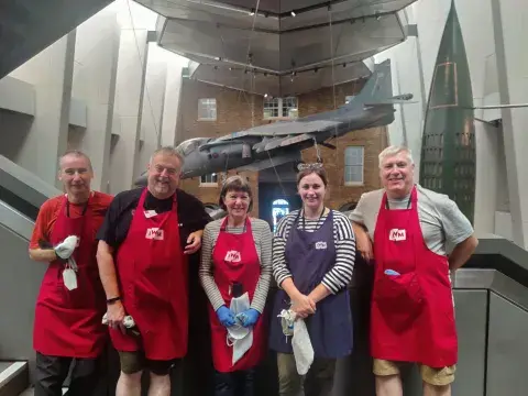 Five smiling preservation volunteers stood on staircase at IWM London, some holding preservation equipment. 