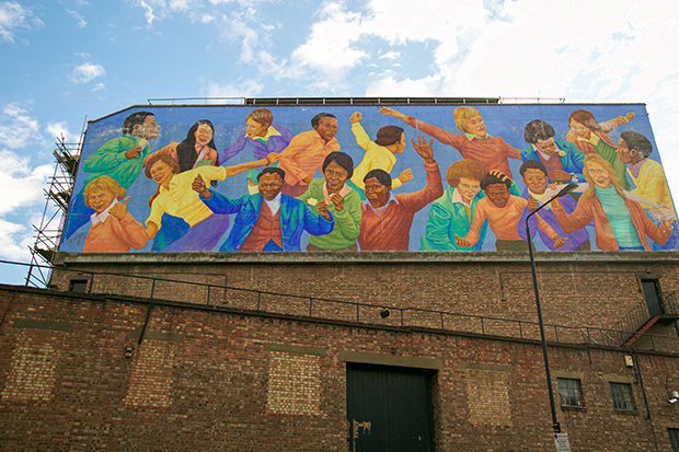 Mural painted on the side of a brick building. The mural depicts 18 children of different races running, playing and holding each other. The background is bright blue and their clothing is colourful: purple, yellow, red and orange.