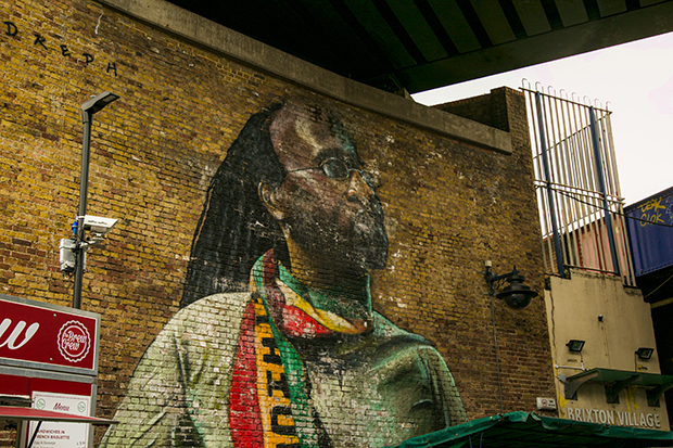 Mural painted on a brick wall beneath a railway bridge. The mural depicts Michael Johns, a local resident. Johns has long dreadlocks and small round glasses. In the painting he looks calmly upward while wearing a green t-shirt and red, yellow and green scarf.